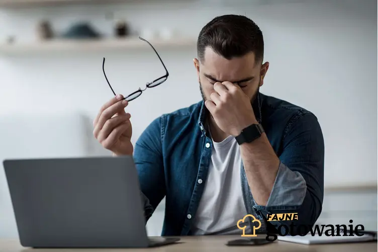 Mężczyzna siedzi przed laptopem, w jednej ręce trzyma okulary, a drugą ręką przeciera oczy, a także które składniki mineralne i witaminy są bardzo ważne w prawidłowym widzeniu.