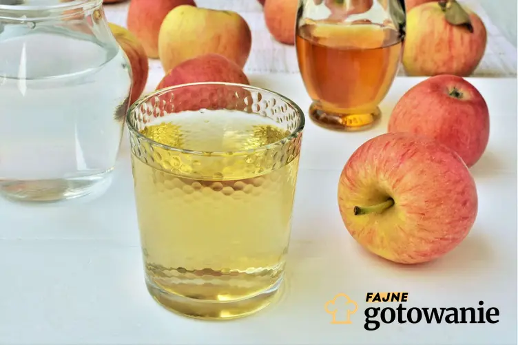 Ocet jabłkowy w szklance, rozcieńczony w wodzie, w tle dzbanek z wodą, ocet jabłkowy i jabłka, a także jak pić ocet jabłkowy