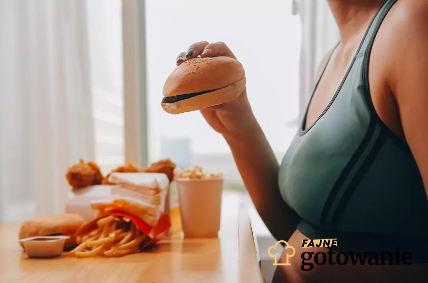 Kompulsywne objadanie wiąże się z wieloma skutkami zdrowotnymi Kobieta trzyma w ręce burgera, przed sobą leży jeszcze więcej fast foodów