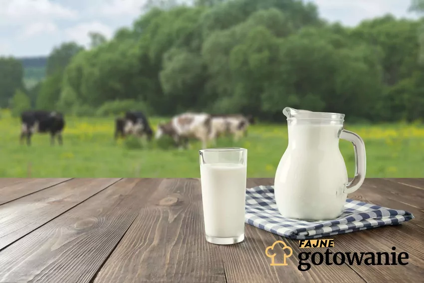 Produkty mleczne to nieodłączny element diety Polaków Dzbanek i szklanka z mlekiem ustawione na stole, w tle łąka z krowami