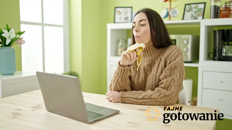 Kobieta siedzi przy stoliku i laptopie i je banana, a także dlaczego nie należy jeść bananów rano
