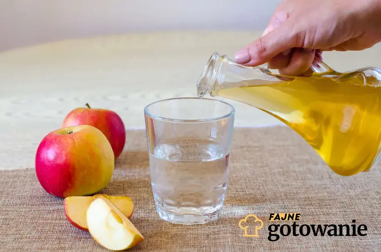 W szklance znajduje się woda, ktoś dolewa octu jabłkowego z dzbanka, w tle są jabłka, a także dlaczego ocet jabłkowy może być szkodliwy