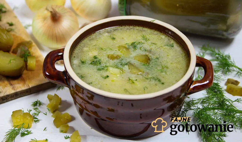 Zupa ogórkowa to jedna z najpyszniejszych zup w kuchni polskiej. Warto znać różne jej warianty Zupa ogórkowa w kamionkowej miseczce. W tle cebula i ogórki kiszone w słoiku