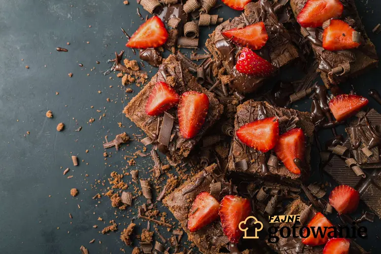 Brownie czekoladowe oblane gorzką czekoladą z położonymi na wierzchu truskawkami pokrojonymi na cztery części