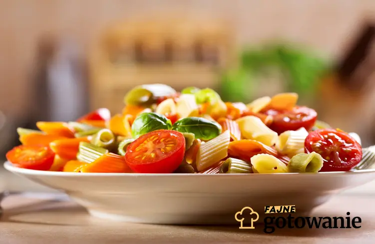 Sałatka z kolorowym makaronem leży na białym talerzu. Widać kolorowy makaron, liście bazylii oraz pomidorki koktajlowe.