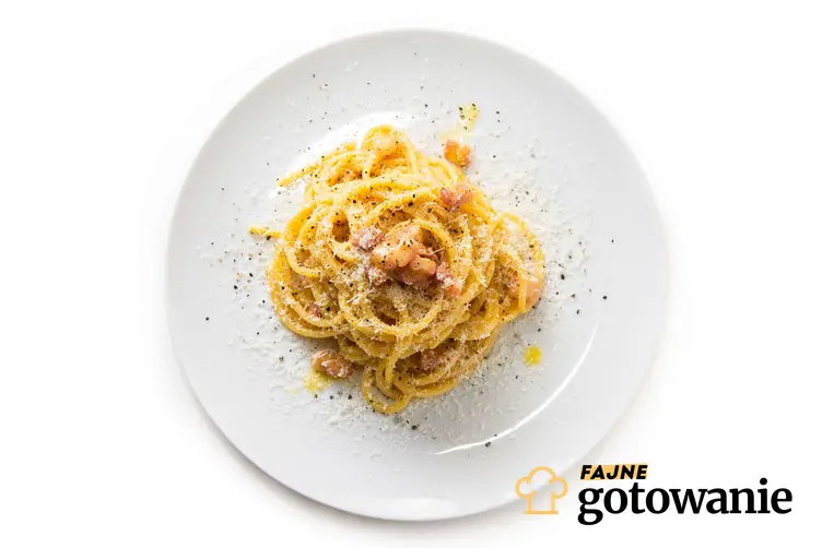 Spaghetti carbonara podane na eleganckim białym talerzu posypane serem i pieprzem.