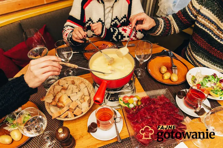 Fondue serowe podane w czerwonym garnuszku na stole. Dookoła stołu siedzą ludzie i maczają pieczywo w fondue.