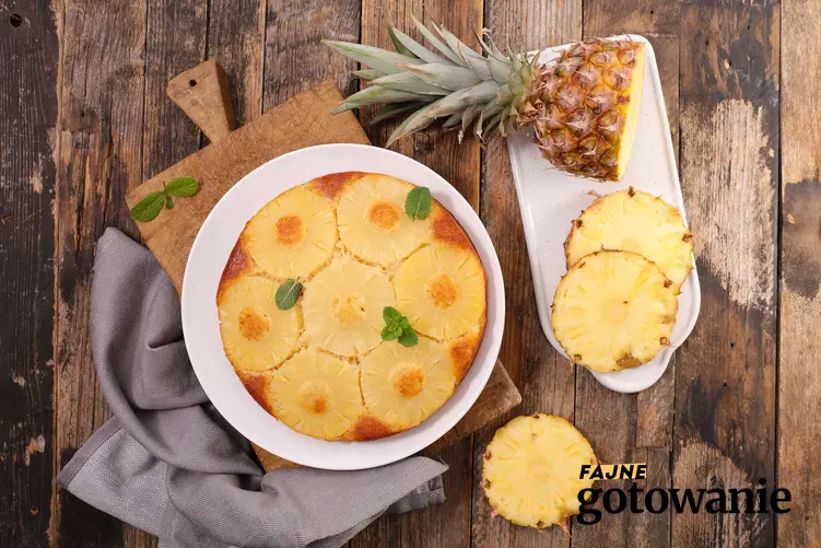 Ciasto z ananasem podane na białym talerzu, na drewnianej desce, obok szara ściereczka i pokrojony świeży ananas, na drewnianym blacie