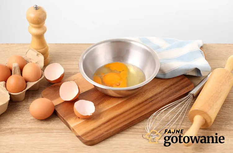 Jajka wbite do srebrnej miski, obok trzepaczka i drewniany wałek.