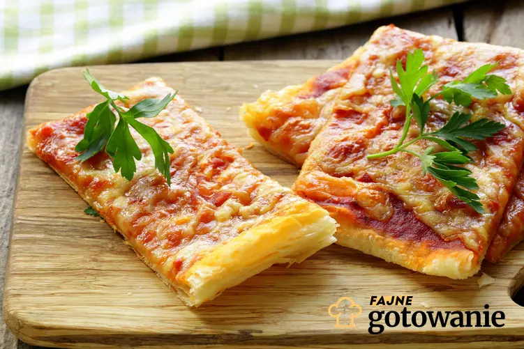 Pizza na cieście francuskim z serem mozzarella i sosem pomidorowym leży pokrojona na drewnianej desce do krojenia. Pizza jest udekorowana natką pietruszki.