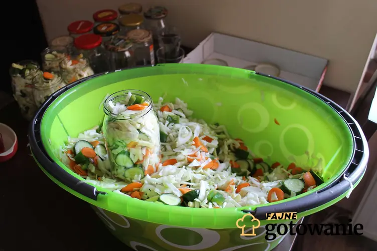 Zielona, plastikowa miska z sałatką wielowarzywną, w której stoi słoik wypełniony warzywami. Za miską znajduje się więcej słoików, zarówno napełnionych warzywami, jak i pustych.