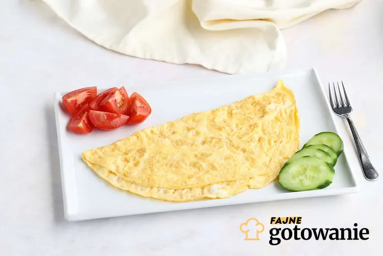 Omlet dla dziecka na białym, prostokątnym talerzu. Obok omletu leży pokrojony pomidor i ogórek.