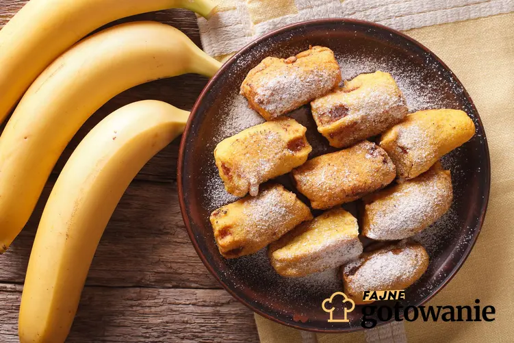 Banan w cieście podany na brązowym talerzu, na kawałku papieru, obok trzy banany a całość przedstawiona na drewnianym stole
