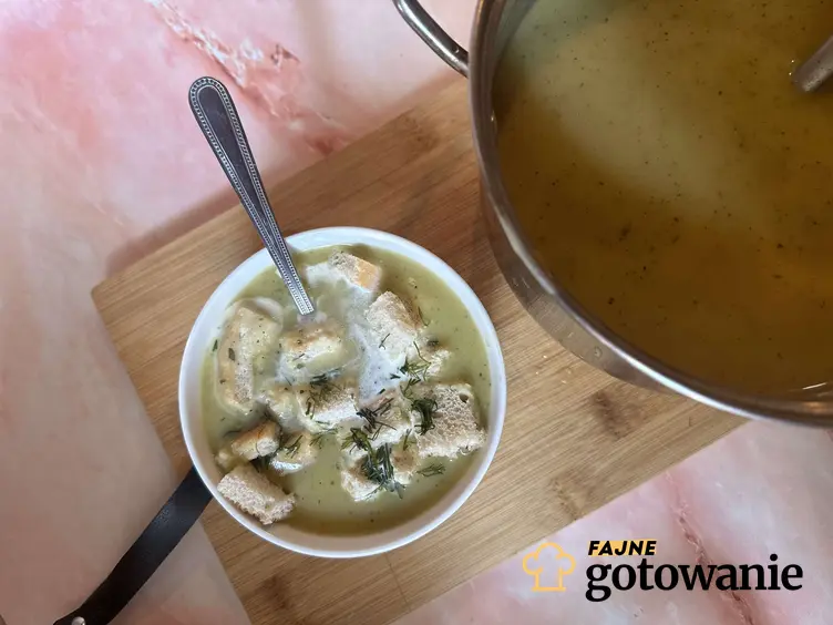 Zupa krem z cukinii z ziemniakami znajduje się w białej misce. Obok postawiona jest druga miska z zupą. W pobliżu leżą: miseczka z grzaneczkami, pomidor, koper i czosnek.