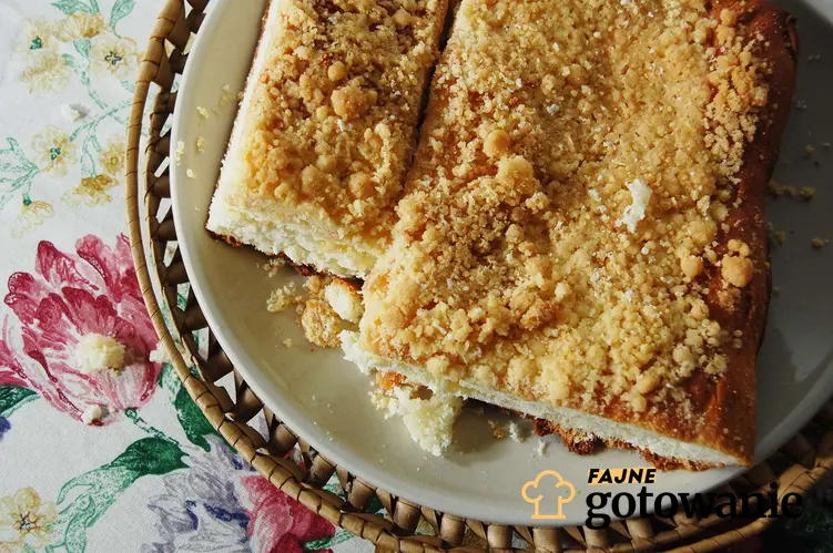 Ciasto drożdżowe z kruszonką, na białym talerzu w wiklinowym koszu leżącym na stole przykrytym obrusem w kwiaty.
