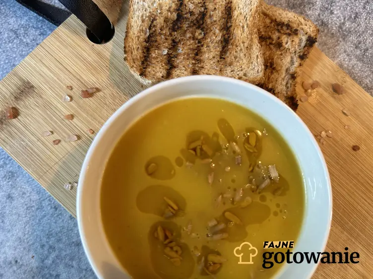 Zupa z kabaczka podana w białej misce, obok grzanki na desce, łyżka i miseczka z pestkami dyni