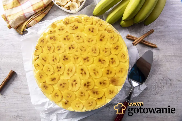 Bananowiec ciasto w okrągłej formie, na białej ściereczce, z łopatką do ciasta, laskami wanilii, bananami i biało-żółtą ściereczką