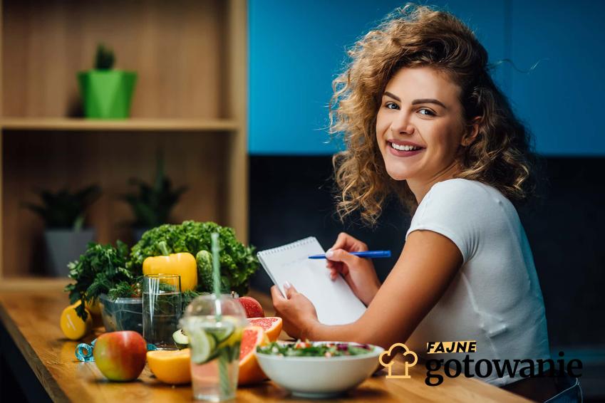 Uśmiechnięta kobieta w kręconych włosach siedzi przy stole na którym stoją miski z owocami i warzywami, jak działa dieta ubogoresztkowa, dieta ubogoresztkowa a picie wody