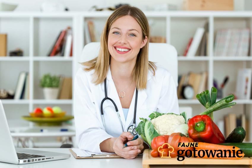 Uśmiechnięta, młoda kobieta w lekarskim uniformie siedzi przy biurku, na którym leży deska z owocami i warzywami, czego nie powinna zawierać dieta osoby po usunięciu woreczka żółciowego