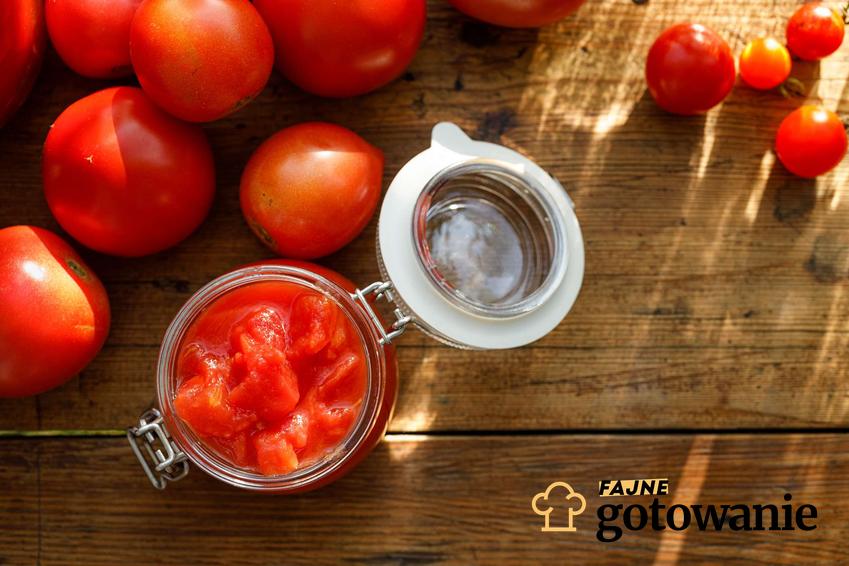 Pomidory w słoikach na zimę podane w słoikach.  Dookoła słoika leżą świeże pomidora.