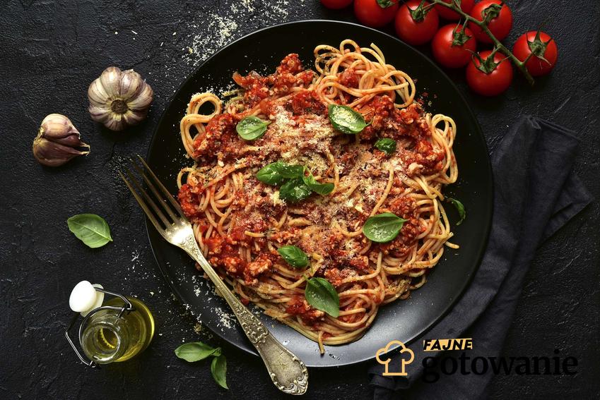 Spaghetti bolognese podane na czarnym talerzu, udekorowane bazylią.