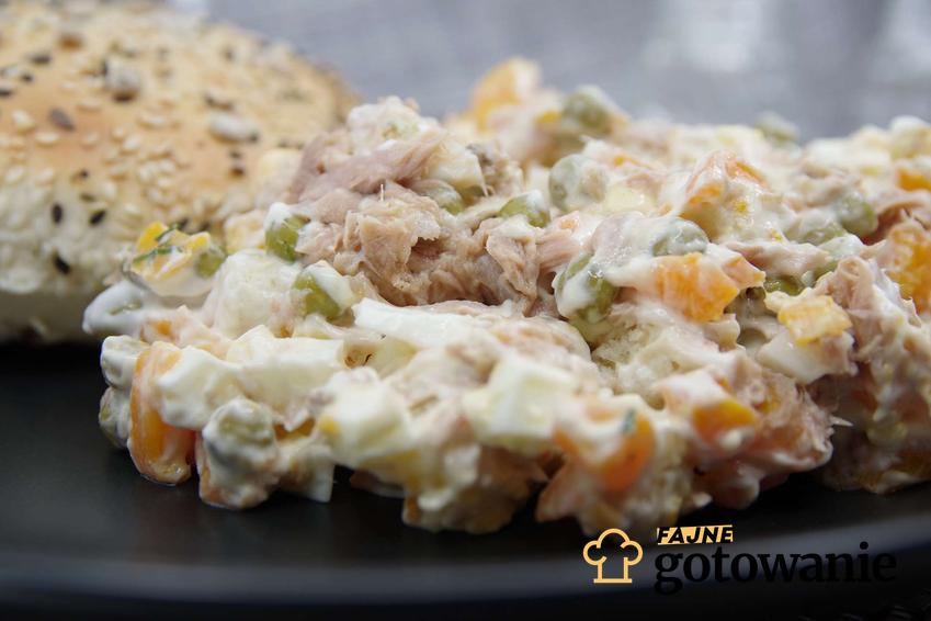 Sałatka z tuńczykiem i kukurydzą, jajkiem, groszkiem i majonezem leży na czarnym talerzu. W tle znajduje się ziarniste pieczywo.
