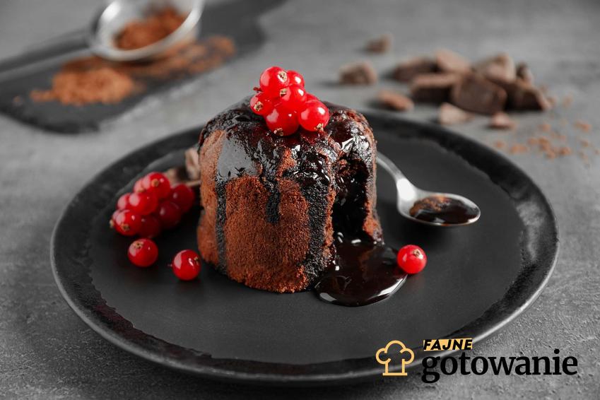 Lava cake podane na czarnym talerzu. Ozdobione jest czerwoną porzeczką.
