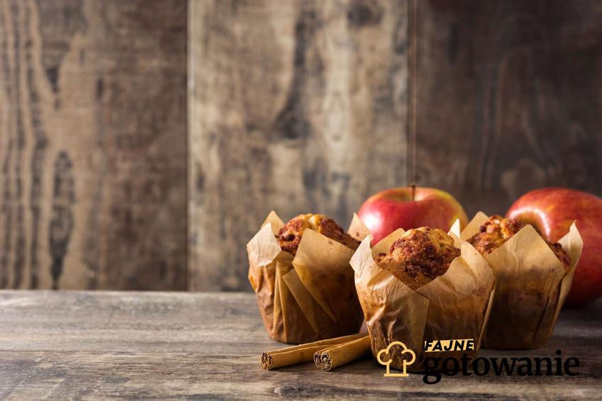 Muffiny z jabłkami ustawione są na stole. Za nimi znajdują się świeże jabłka.