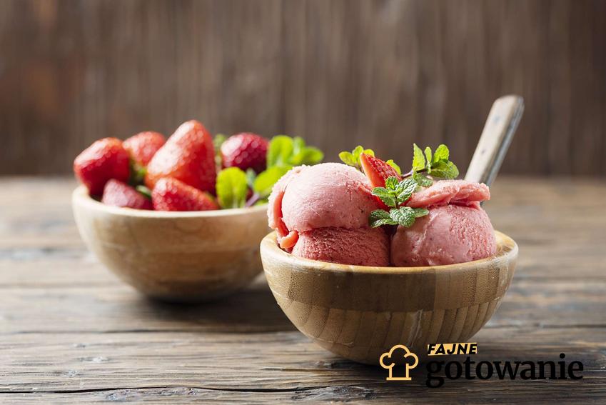 Lody jogurtowe znajdują się w drewnianej miseczce. Lody udekorowane są świeżymi truskawkami i listkami mięty. Obok stoi miseczka z truskawkami i miętą.