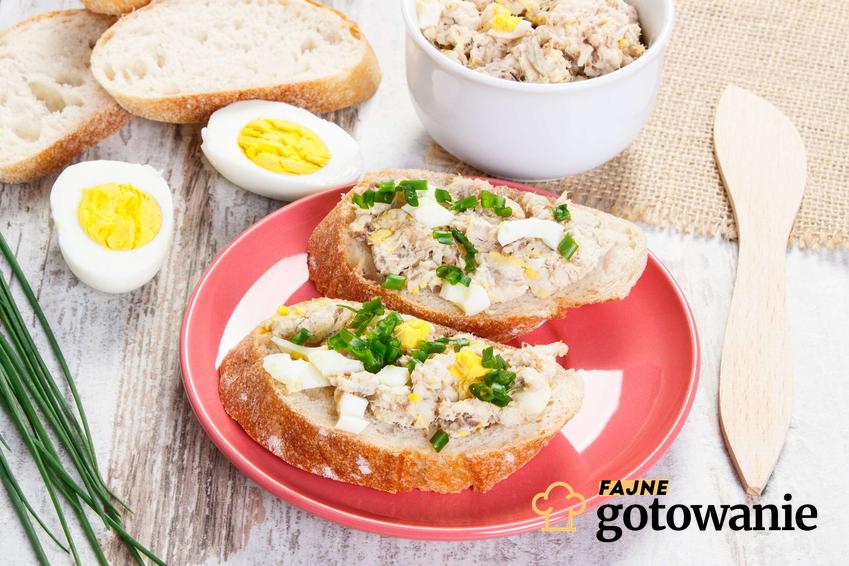 Pasta z makreli i jajek obsypana szczypiorkiem znajduje się na dwóch kromkach chleba. Chleb leży na czerwonym talerzu. Obok znajdują się: przekrojone jajko, miska z pastą, drewniany nożyk i szczypiorek.