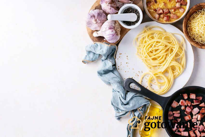 Sos carbonara w trakcie przygotowania, na białym talerzu makaron spaghetti, w miseczkach ser, jajka, czosnek, na patelni żeliwnej podsmażony boczek.