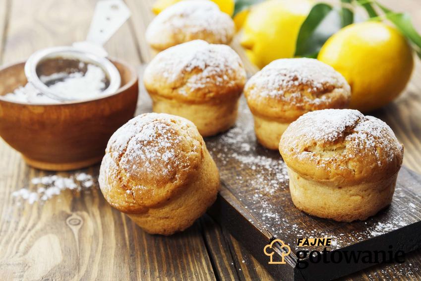 Muffinki cytrynowe posypane cukrem pudrem ułożone są na drewnianym blacie. Leżą na nim również cytryny.