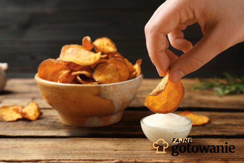 Chips paprykowy maczany w białym, czosnkowym sosie. Z tyłu, na drewnianym stole, stoi miseczka z chipsami.