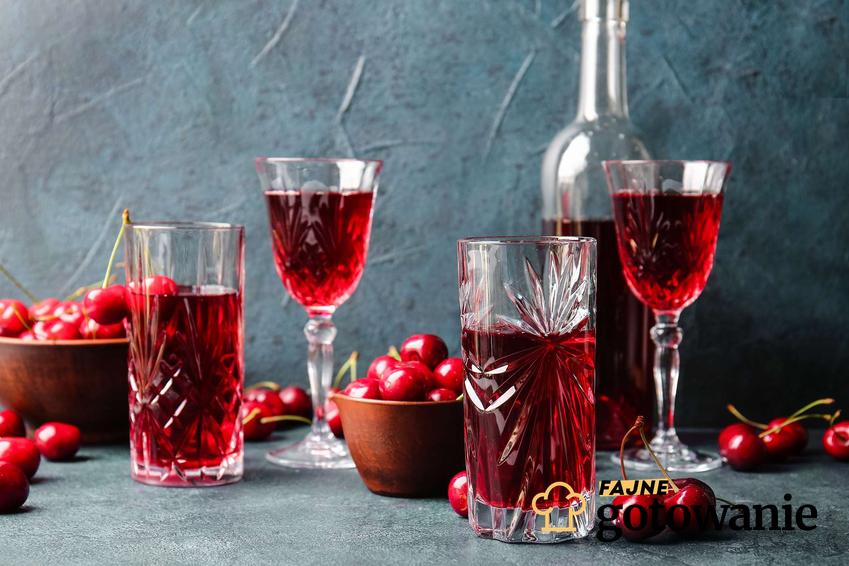 Wino z wiśni podane w kryształowych szklankach i lampkach, z tyłu butelka z winem oraz miseczki z wiśniami.