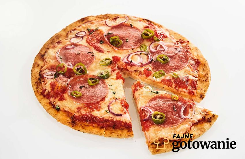 Diavolo pizza z salami, ostrą papryczką i czerwoną cebulką leży na białym blacie.