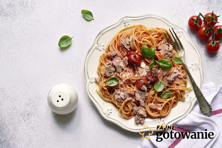 Spaghetti z tuńczykiem podane na białym, ozdobnym talerzu. Dookoła talerza znajdują się listki bazylii oraz pomidorki koktajlowe.