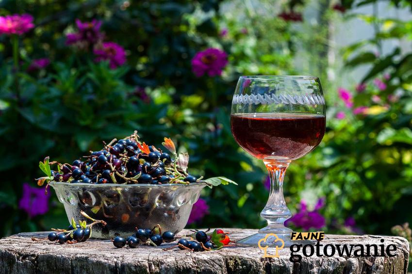 Wino z czarnej porzeczki podane jest w kieliszku obok którego znajduje się miseczka ze świeżymi owocami czarnej porzeczki.