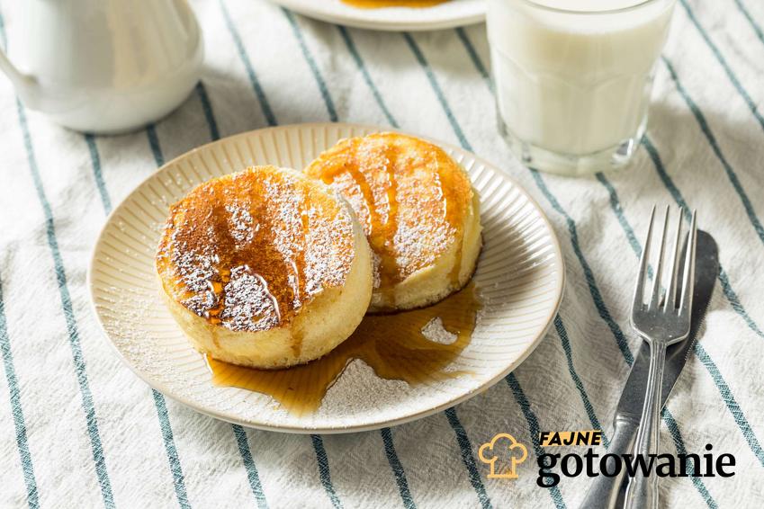 Japońskie pancakes polane syropem klonowym i oprószone cukrem pudrem leżą na białym talerzyku. Obok leżą sztućce i szklanka z mlekiem.