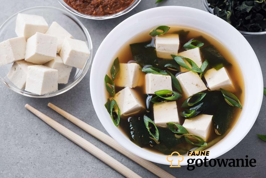 Zupa miso z tofu w białej miseczce. Obok pałeczki oraz tofu i wodorosty wakame.