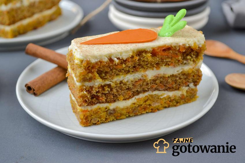 Ciasto marchewkowe z mascarpone podane na białym talerzu z dwoma laskami cynamonu, na kawałku ciasta marchewka z masy cukrowej