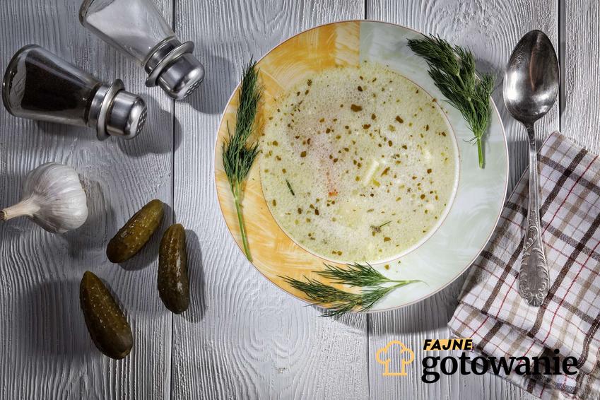Zupa ogórkowa na żeberkach znajduje się w biało-żółtym talerzu udekorowanym koperkiem. Obok leżą ogórki, ściereczka, czosnek, łyżka, solniczka i pieprzniczka.