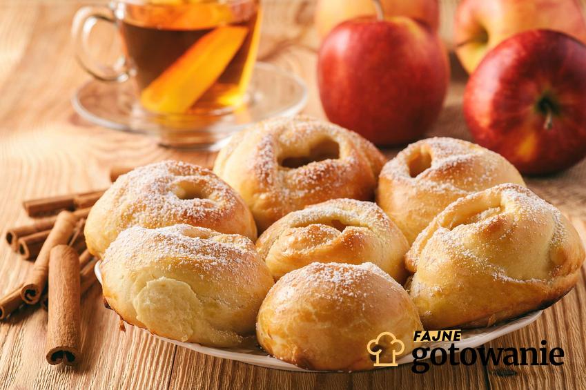 Bułki drożdżowe z jabłkami podane na talerzyku, obok laski cynamonu, szklanka z herbatą, świeże jabłka