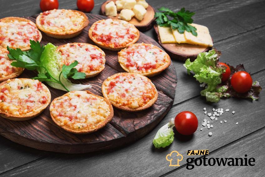 Mini pizza (9 sztuk) znajduje się na drewnianej deseczce. Obok porozkładane są dodatki na pizzę: ser mozzarella, pomidorki koktajlowe i sałata.