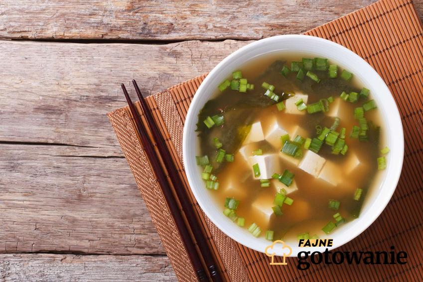 Zupa miso z tofu znajduje się w wysokiej białej misce. Oboj niej znajdują się pałeczki. Miska stoi na brązowej macie bambusowej leżącej na drewnianym blacie.