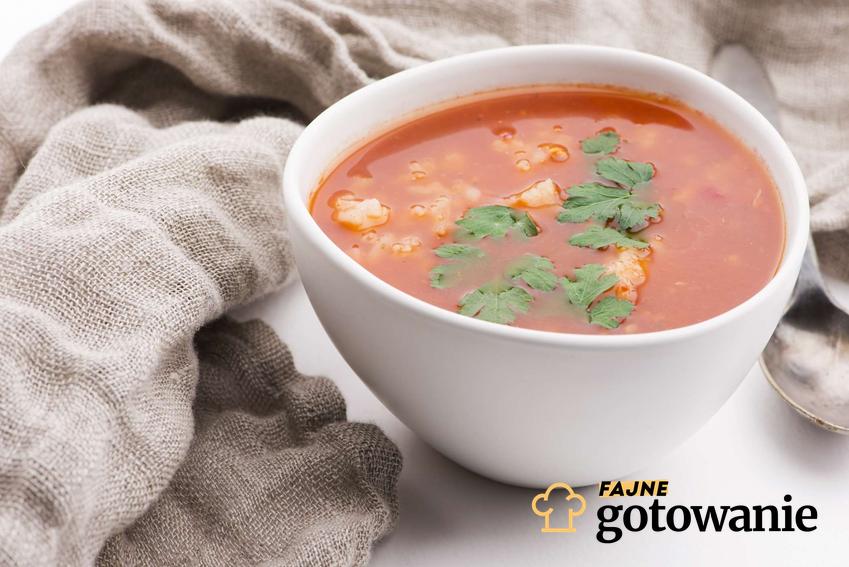 W białej bulionówce znajduje się zupa pomidorowa z ryżem, udekorowana listkami pietruszki. Obok miseczki leży łyżka oraz materiał w jasnym kolorze.