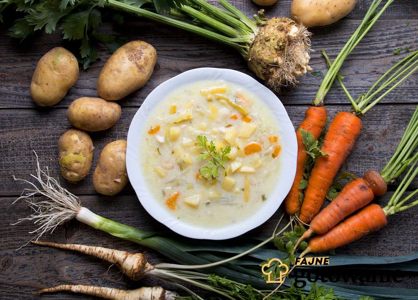 Zupa jarzynowa w białym głębokim talerzu otoczona świeżymi warzywami.