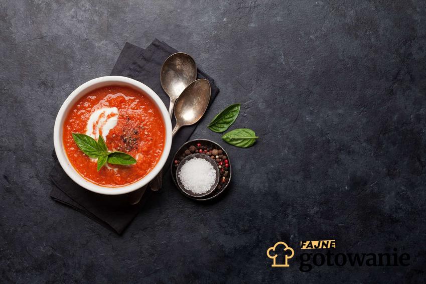 Zupa pomidorowa z passaty podana w białej miseczce. Obok niej znajdują się małe miseczki z przyprawami.