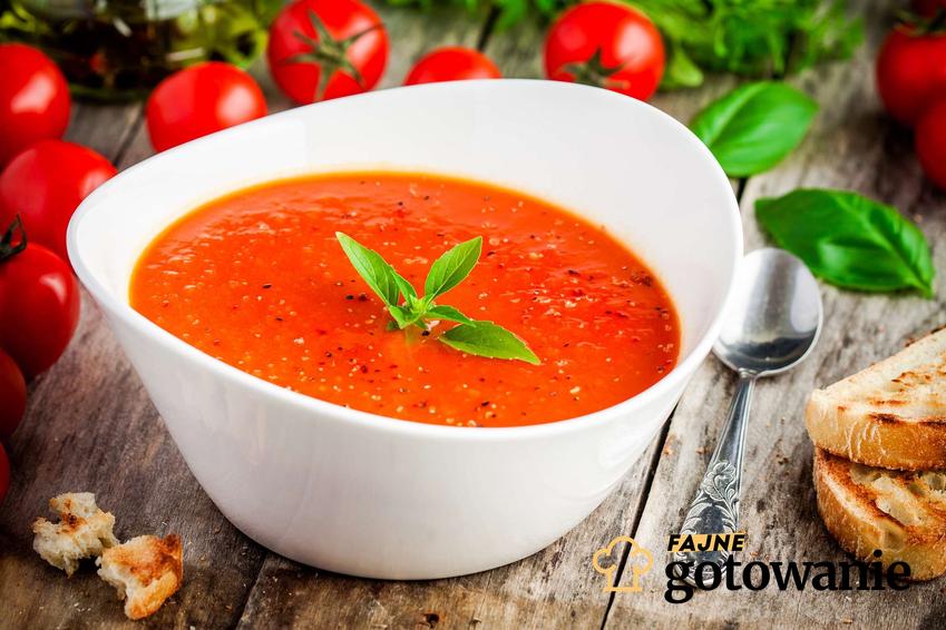 Zupa pomidorowa bez mięsa podana w białej miseczce. Dookoła leżą świeże pomidory i metalowa łyżeczka.