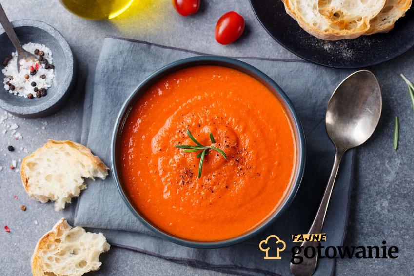 Zupa paprykowa podana w szarej miseczce, na szarej ściereczce, na szarym blacie. Obok łyżka, pomidorki, chleb, oliwa i przyprawy w miseczce.