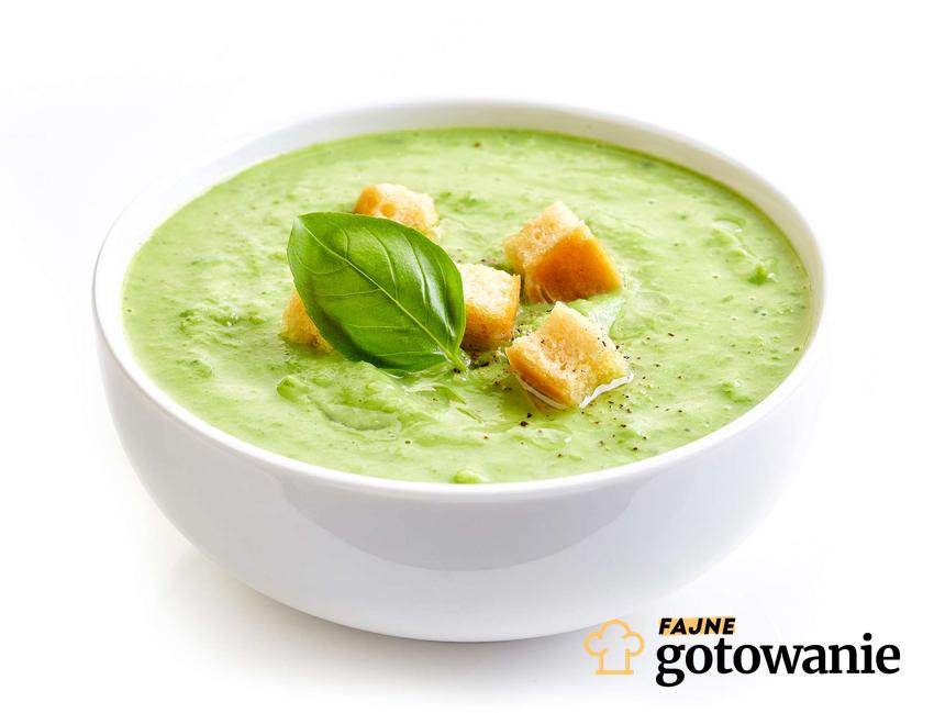 Zielona zupa krem w białym naczyniu, podana z grzankami i udekorowana listkami bazylii.